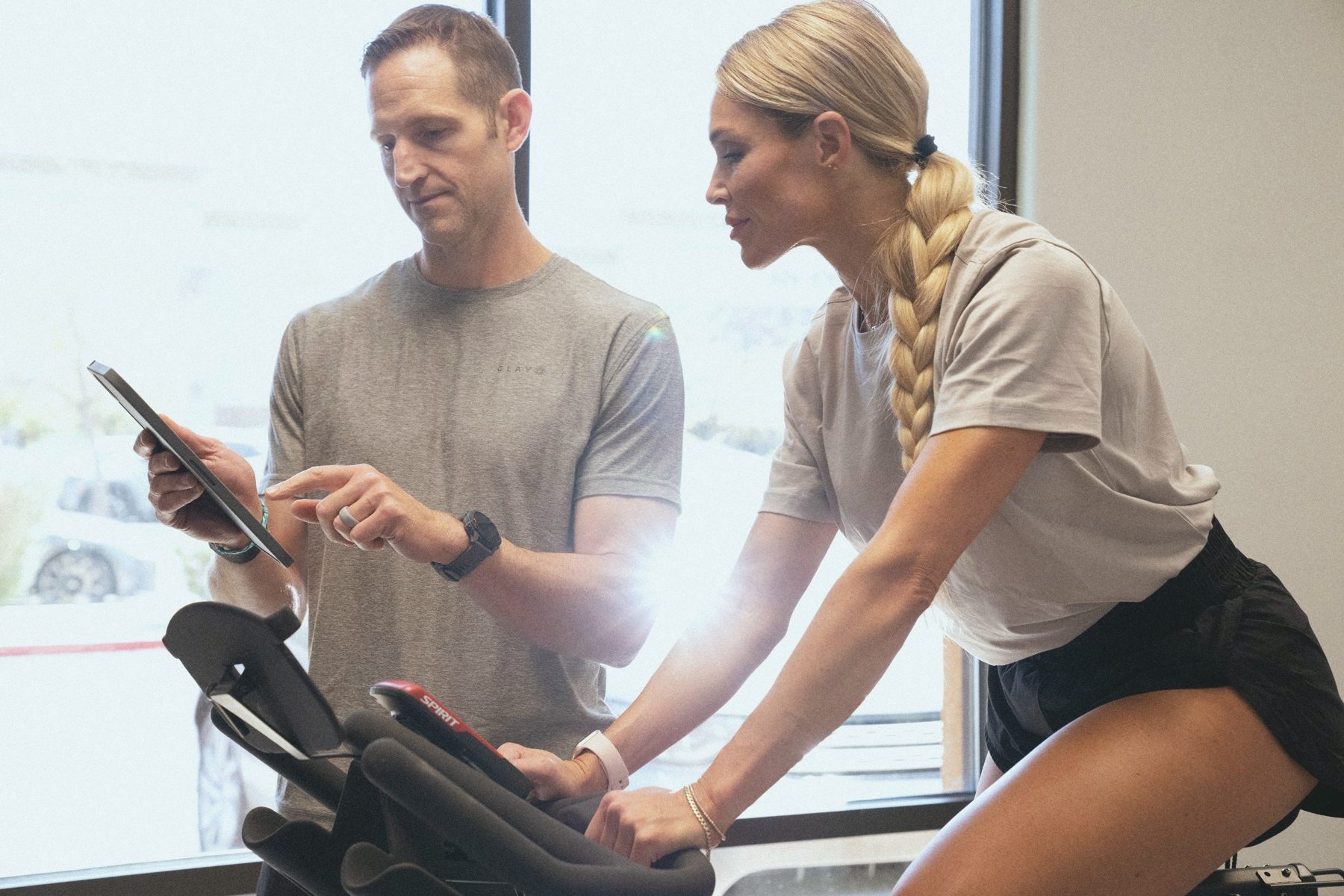A team member reviews a test on a tablet beside a member on an exercise bike.